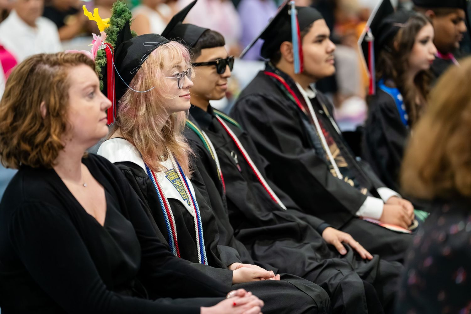 Graduates in black caps and gowns seated at a graduation ceremony, listening attentively to speakers on stage.
