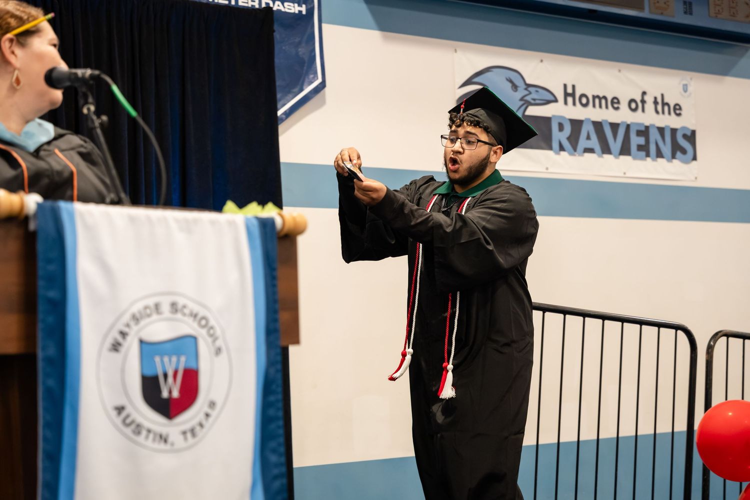 Graduate in cap and gown signing at Wayne School Austin Texas Ravens graduation ceremony on stage.