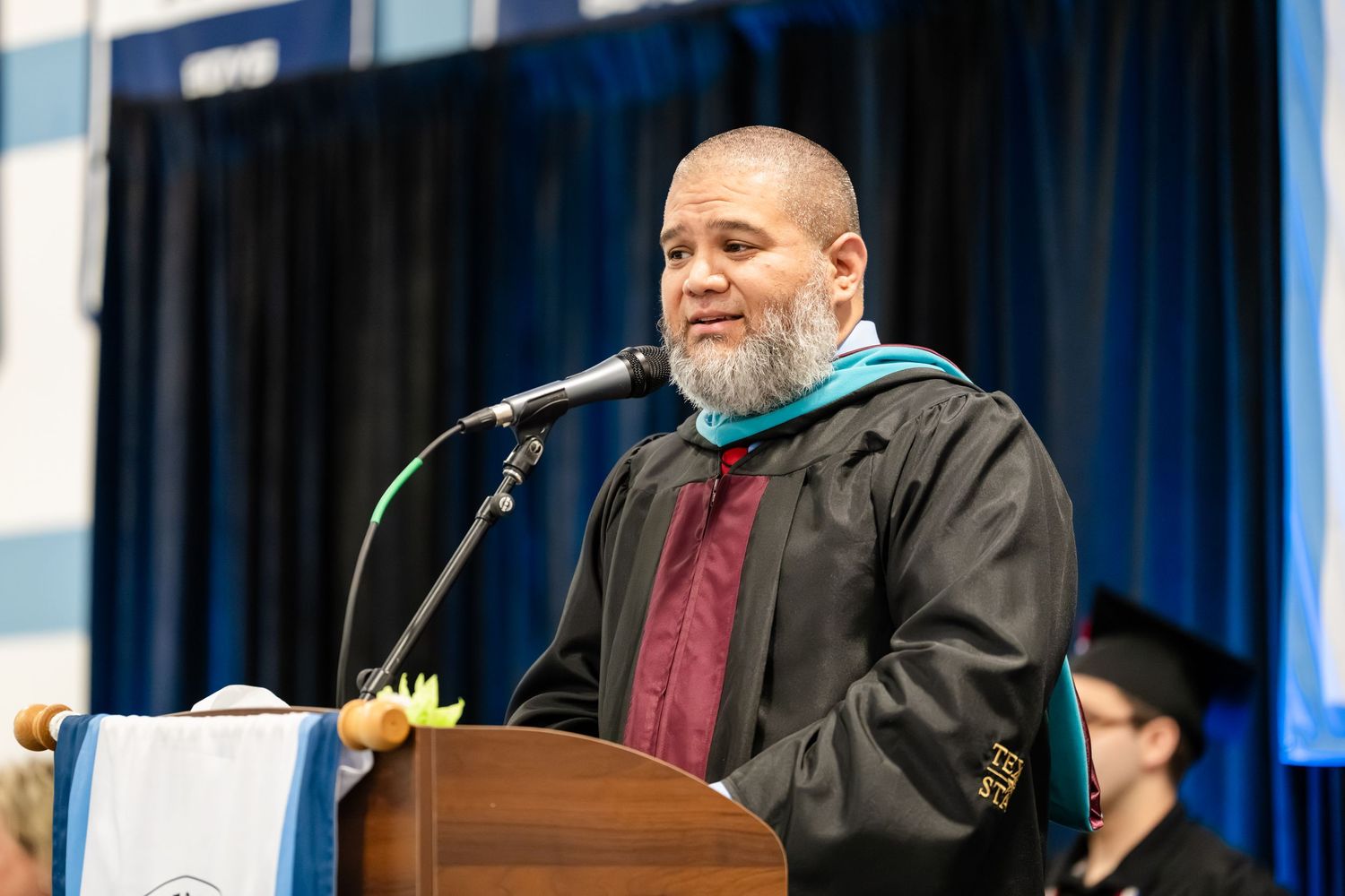 Man in graduation regalia speaking at podium during commencement ceremony.