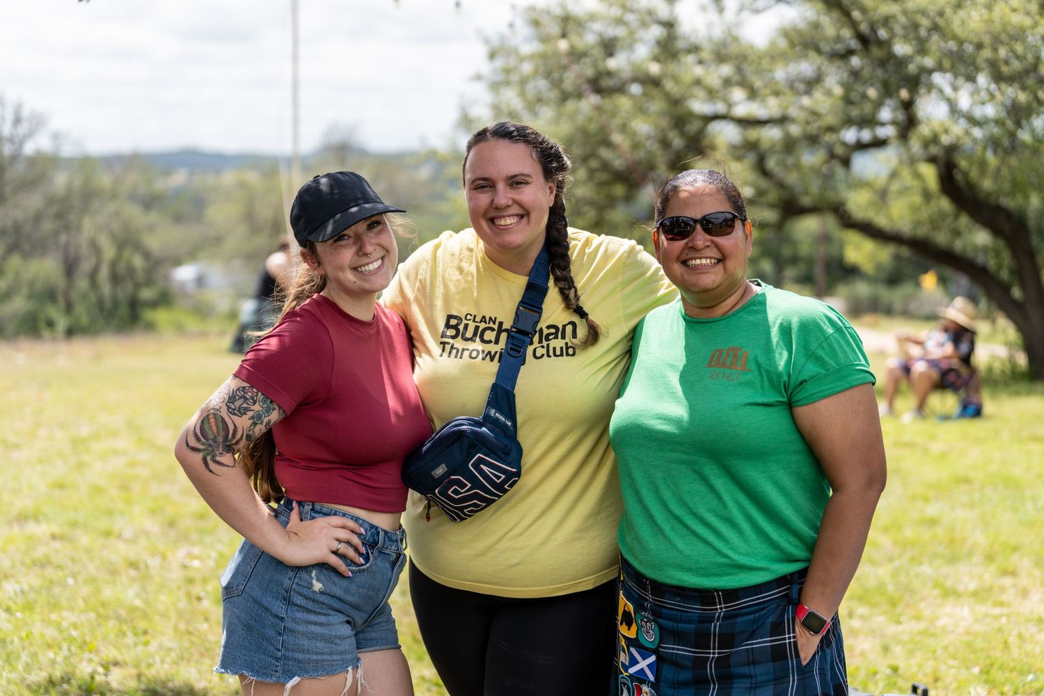 Three women smiling together outdoors at a sunny festival, wearing casual summer clothes on green grass.