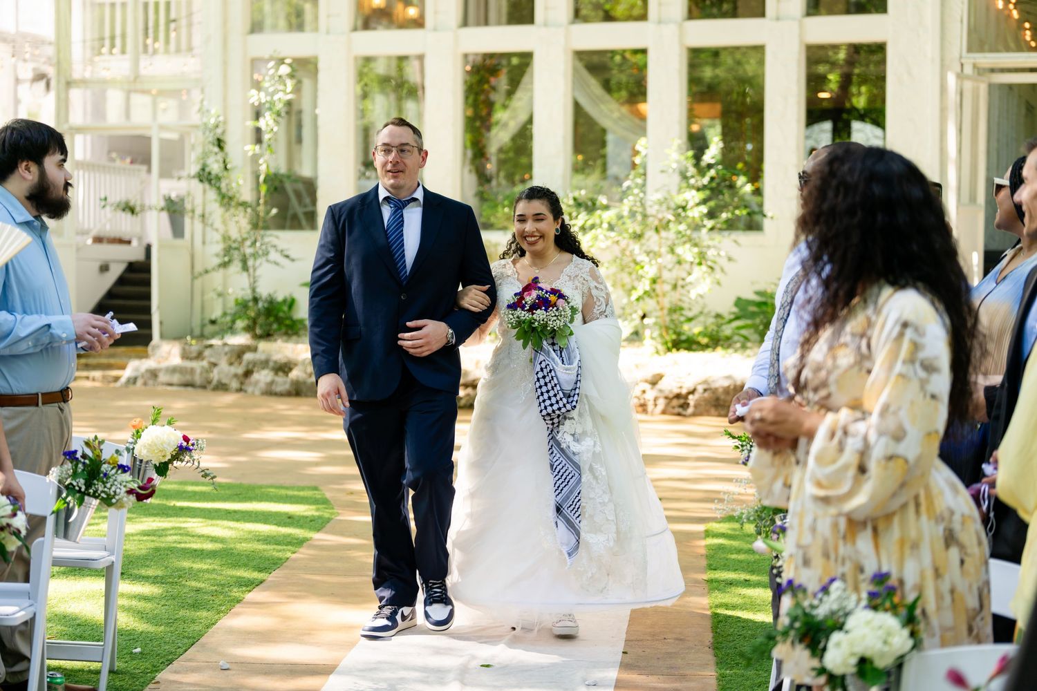 Bride walks down aisle with escort at outdoor wedding ceremony in lush garden venue.