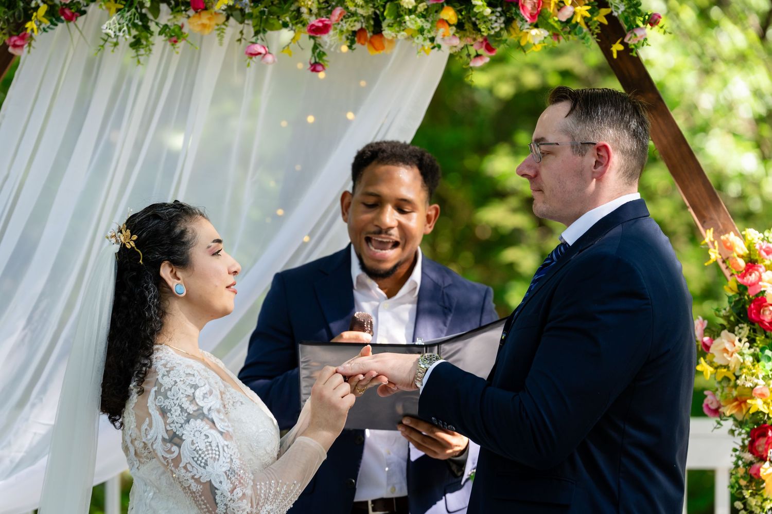 Bride and groom exchanging vows under floral arch with smiling officiant at outdoor wedding ceremony.