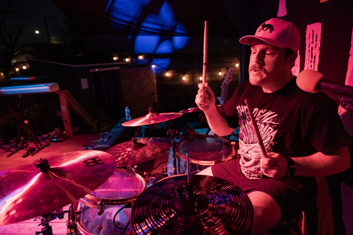 A drummer performs energetically on stage under red lighting, holding drumsticks above a cymbal-filled kit.