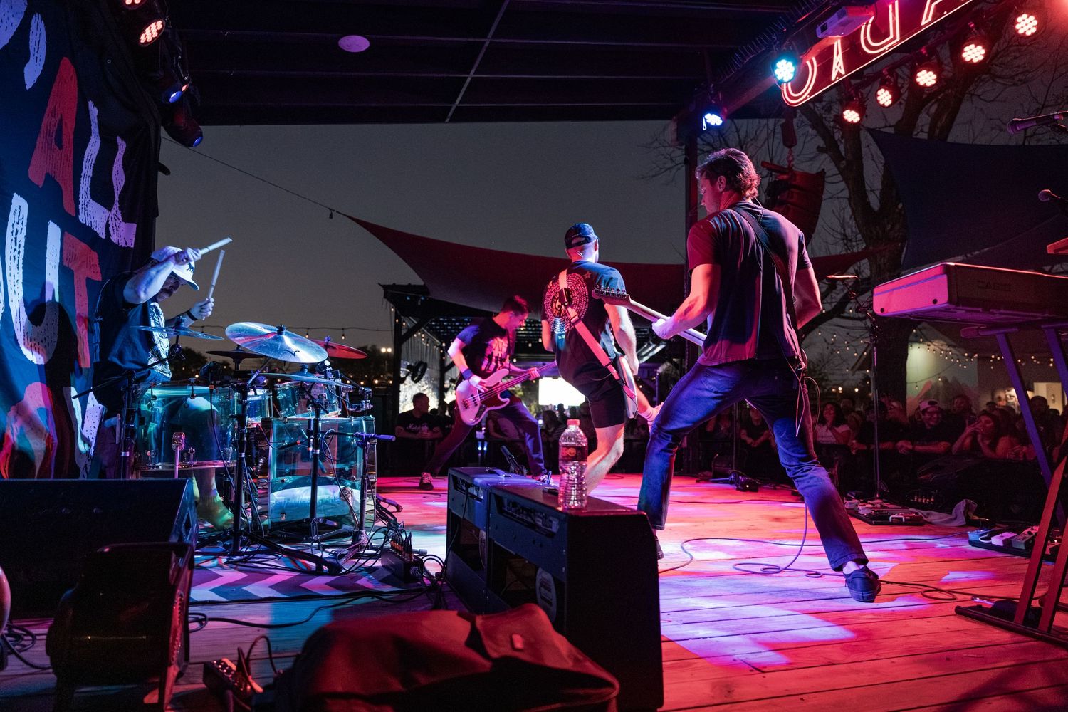 Live rock band performing on an outdoor stage at night with colorful lights illuminating the energetic musicians.