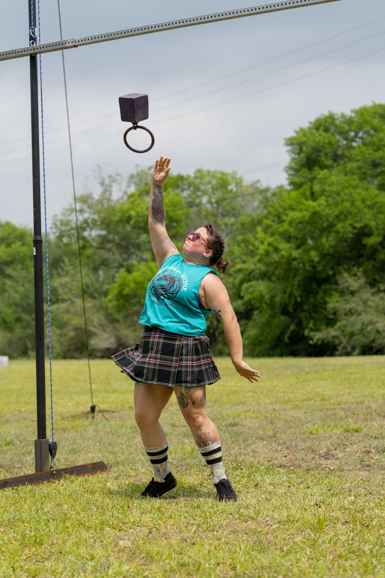 Athlete in tartan skirt and teal tank top competes in Highland Games weight toss on grassy field.