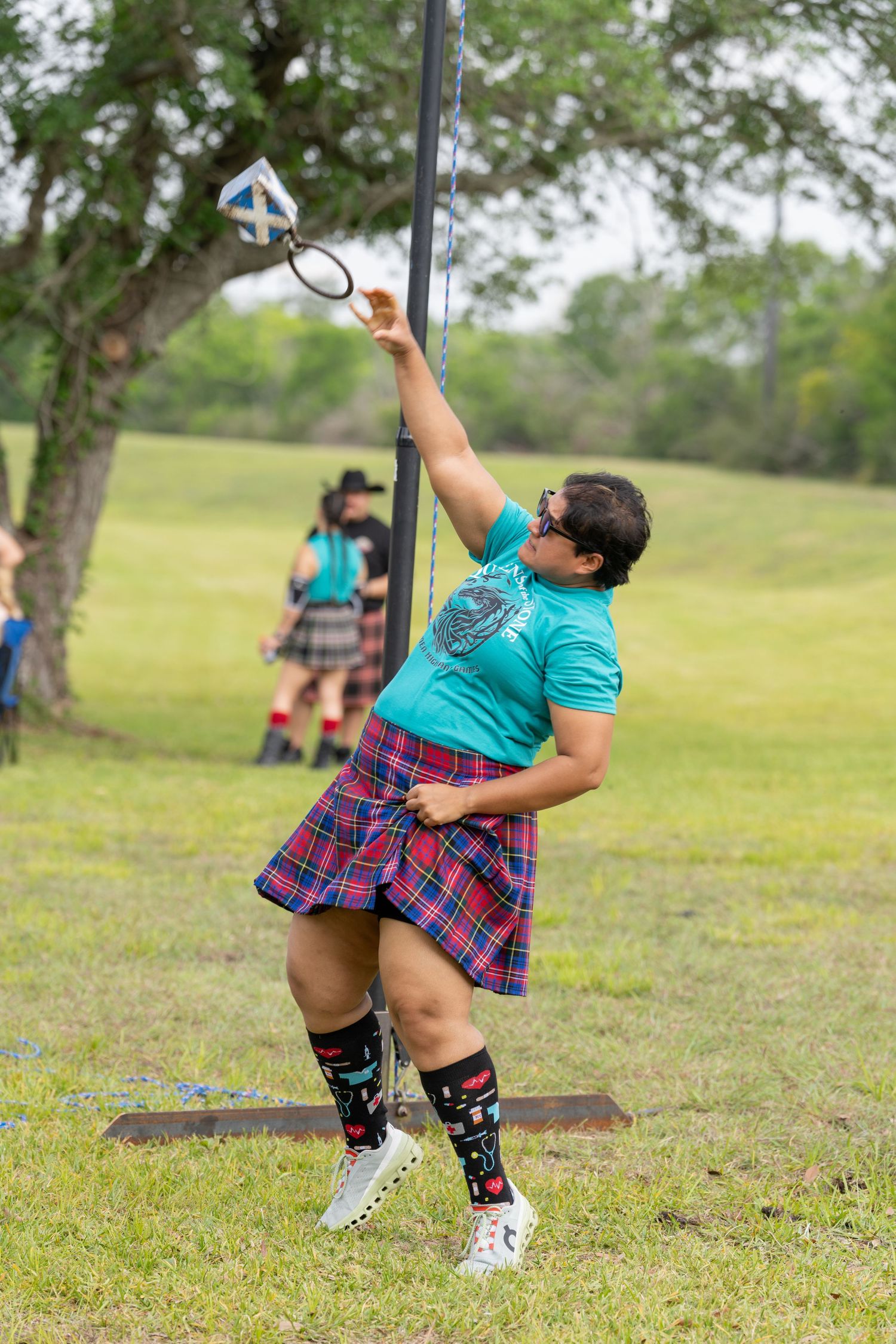 Woman in tartan kilt and teal shirt competing in Highland Games weight-over-bar event on green field.