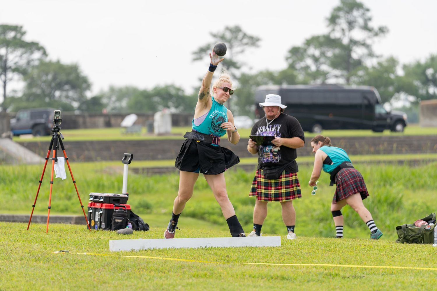 Athletes compete in Highland Games throwing event on grass field, wearing kilts and traditional attire at outdoor festival.