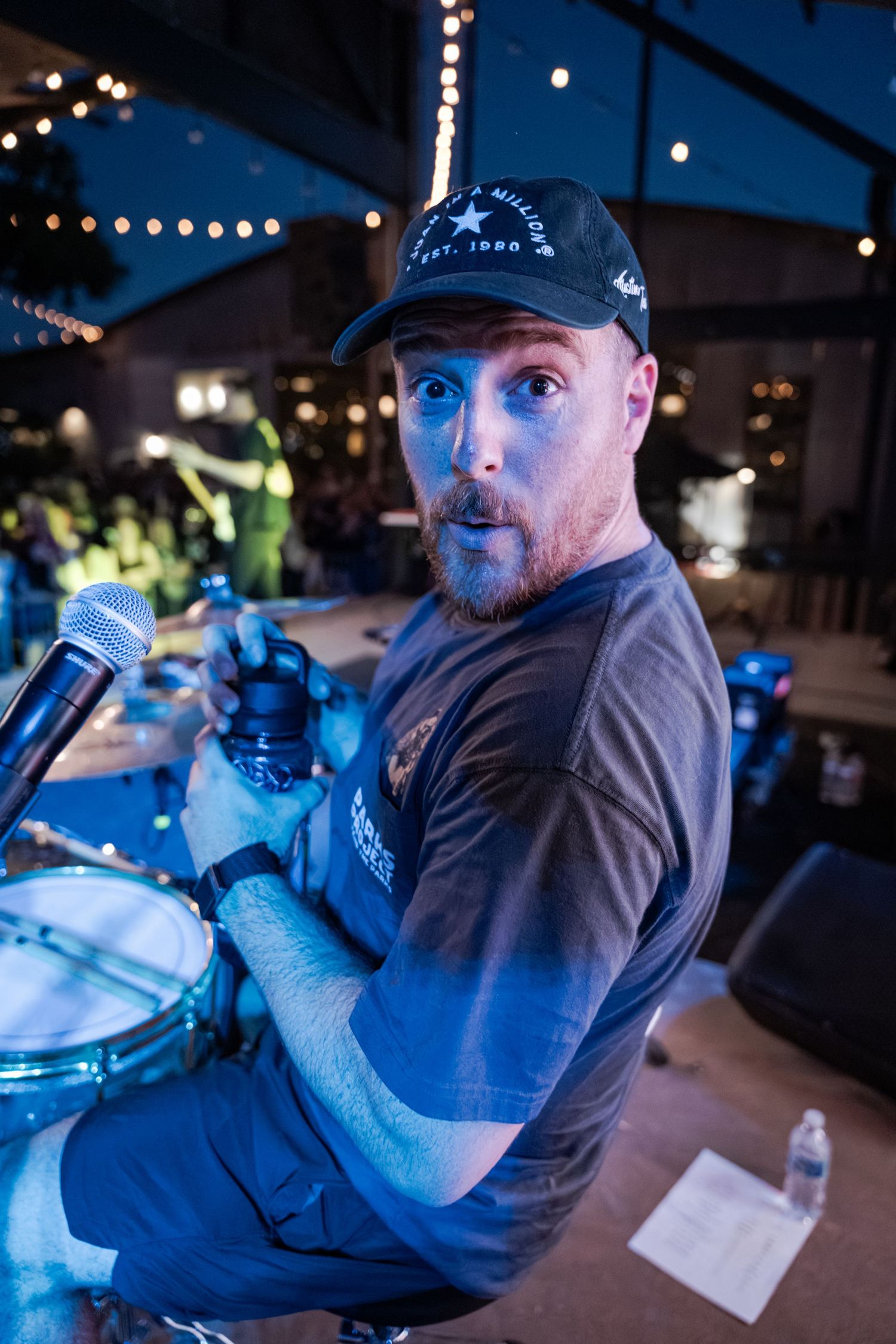 Bearded man in black cap plays drums on stage under string lights at an outdoor music venue.