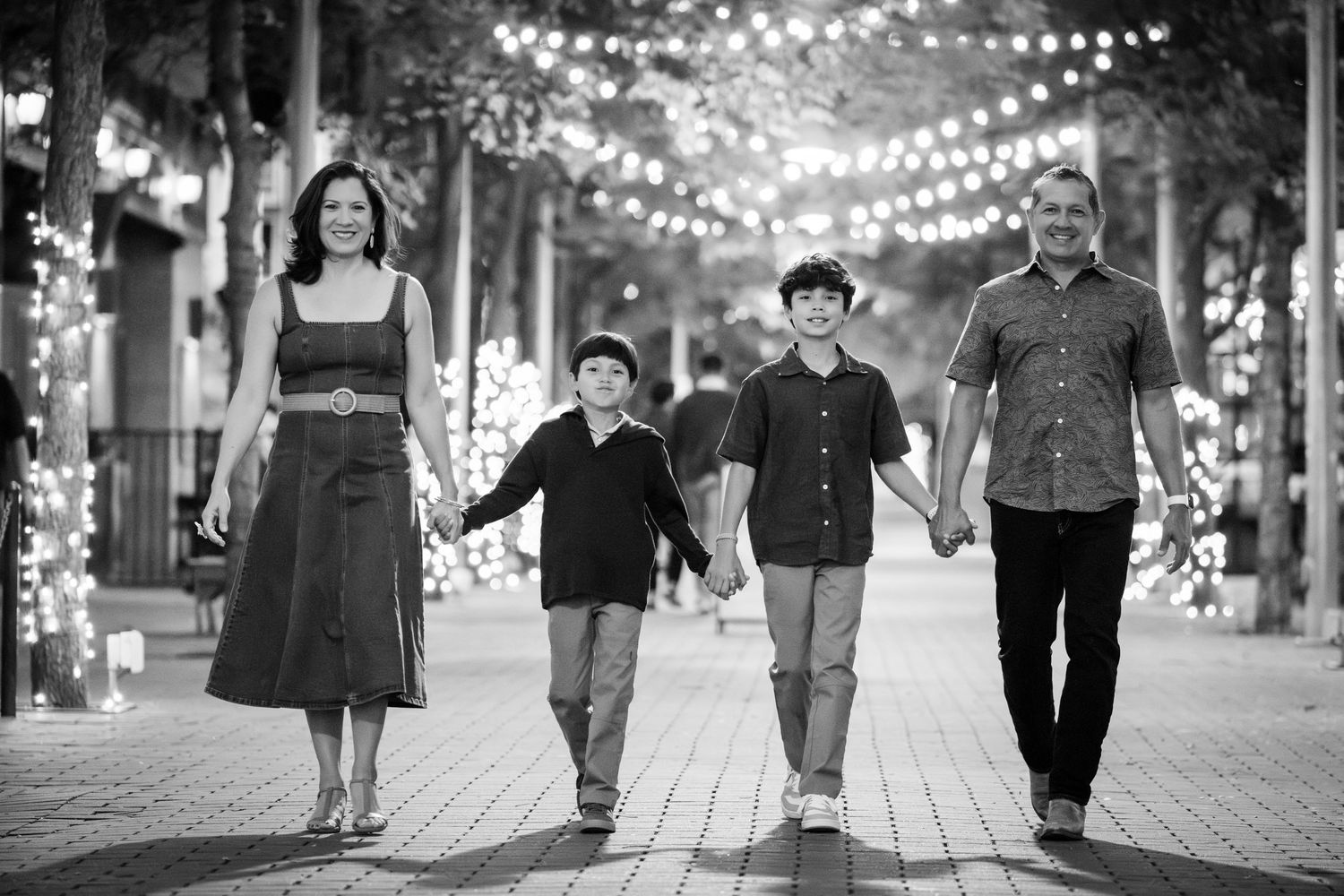 Black and white family photo of parents and two boys walking hand in hand on a festive lit pathway.