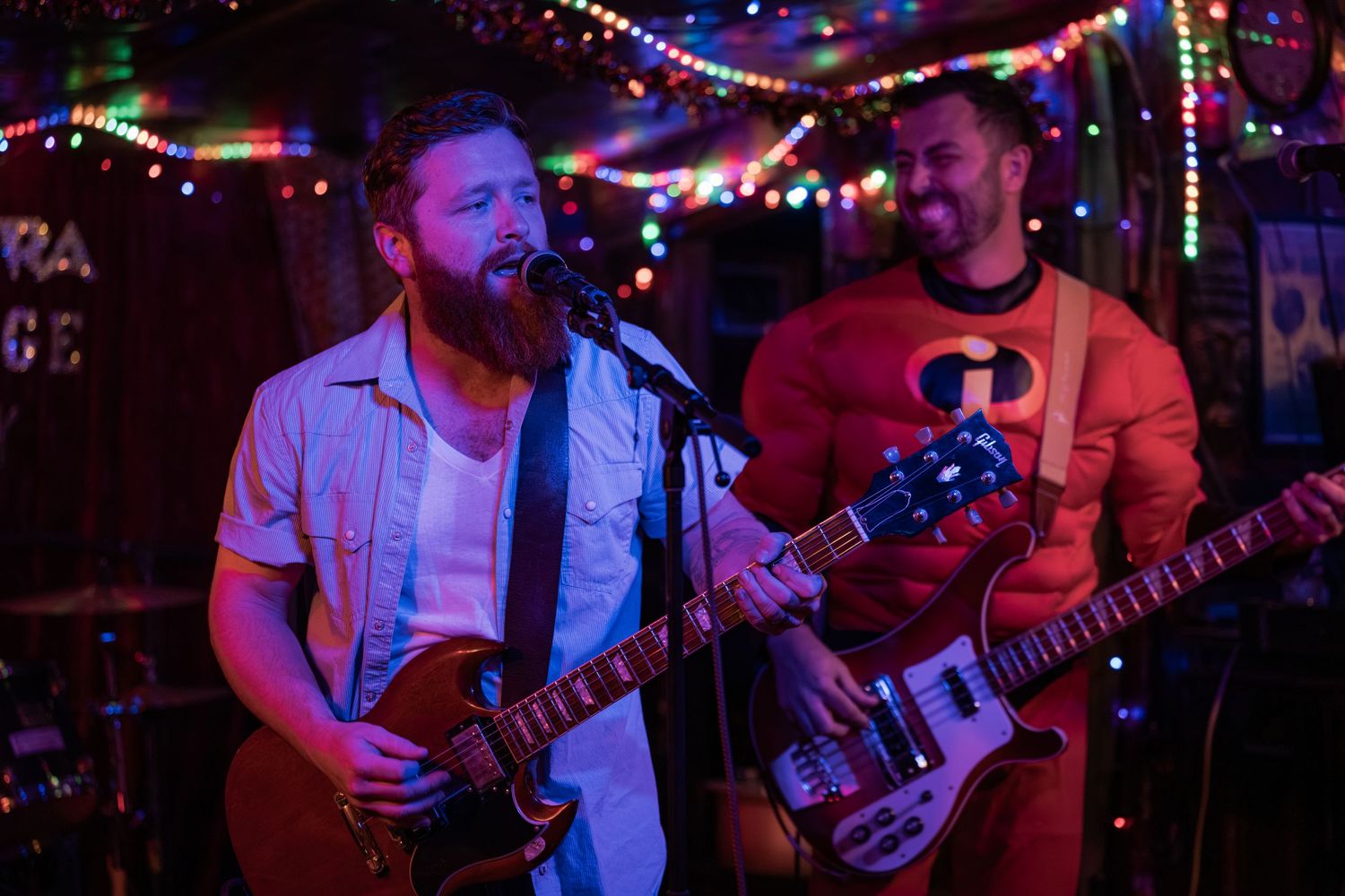 Musicians perform on stage at a dimly lit venue with colorful string lights creating an intimate atmosphere.