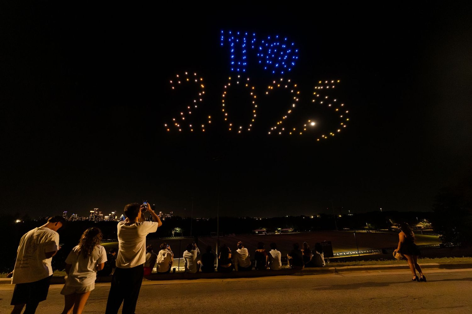 Crowd watches illuminated drone display forming '2025' in blue and red lights against dark night sky.