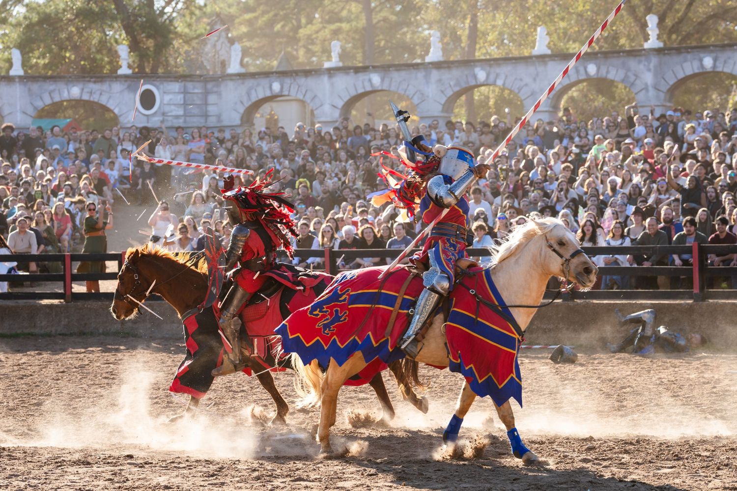 Medieval knights joust on horseback at a Renaissance festival with stone arches and crowd in background.