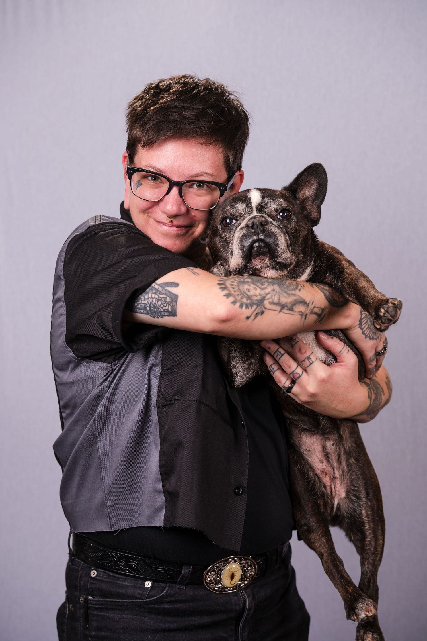 A series of four photos showing someone in a dark vest posing playfully with a French Bulldog against a gray background.