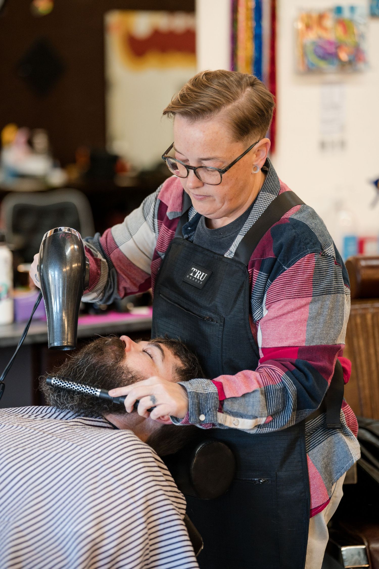 A barber wearing a black vest and pink plaid shirt trims a client's hair with precision in a barbershop setting.