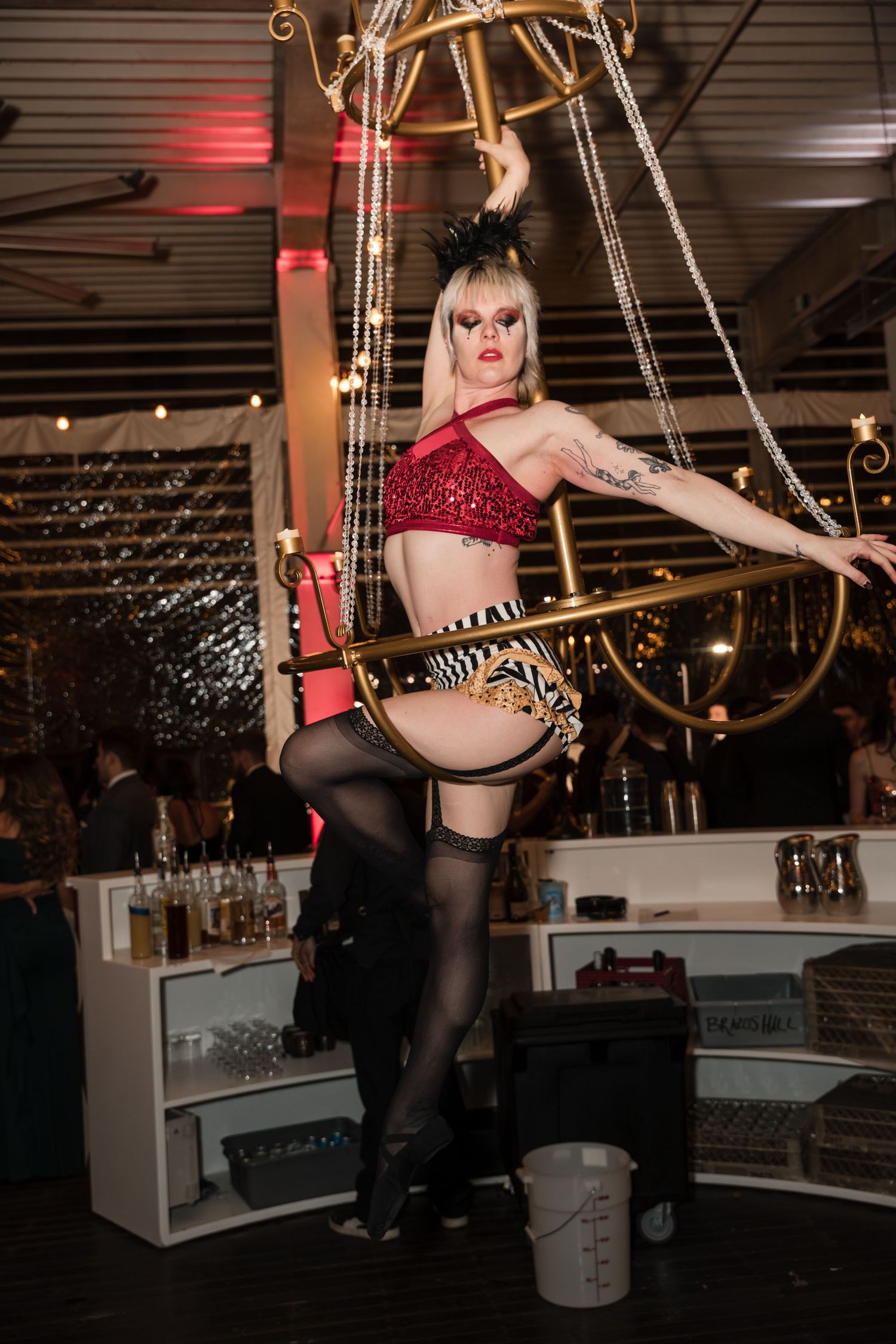 A performer in a red sequined outfit poses dramatically on a swing in a dimly lit venue with chandeliers.