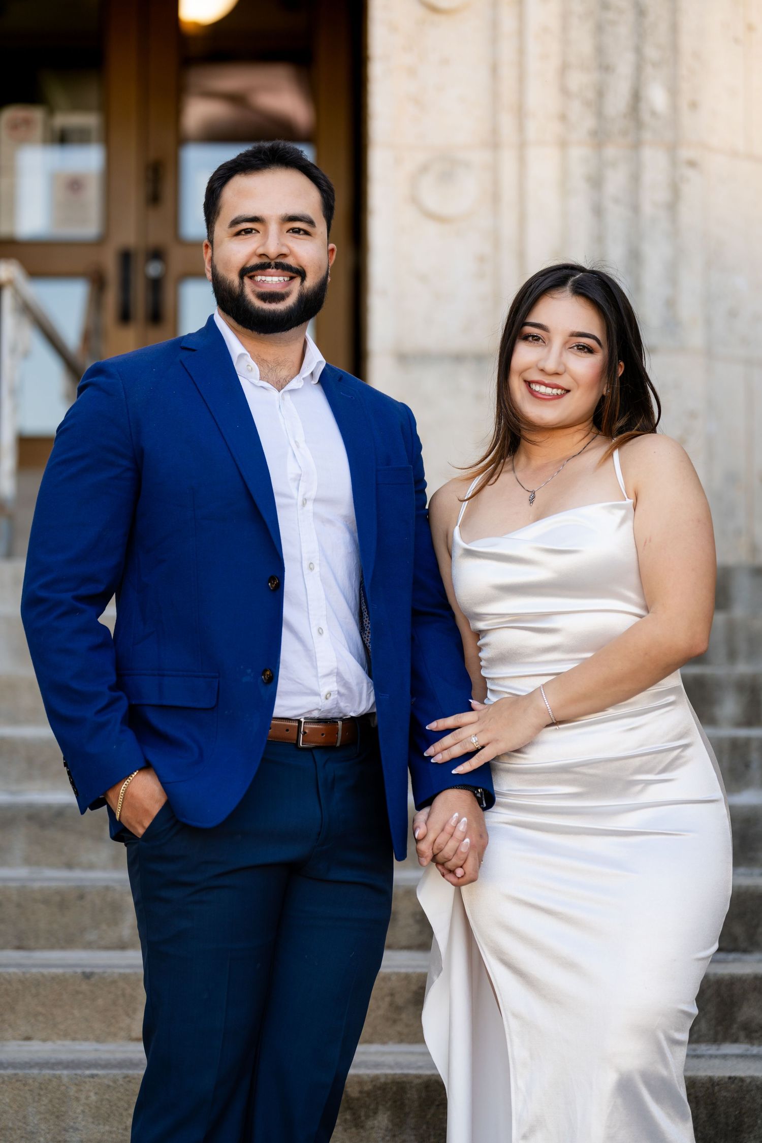 A couple in formal attire poses on stone steps, with the person in a navy suit and white shirt standing next to their partner.