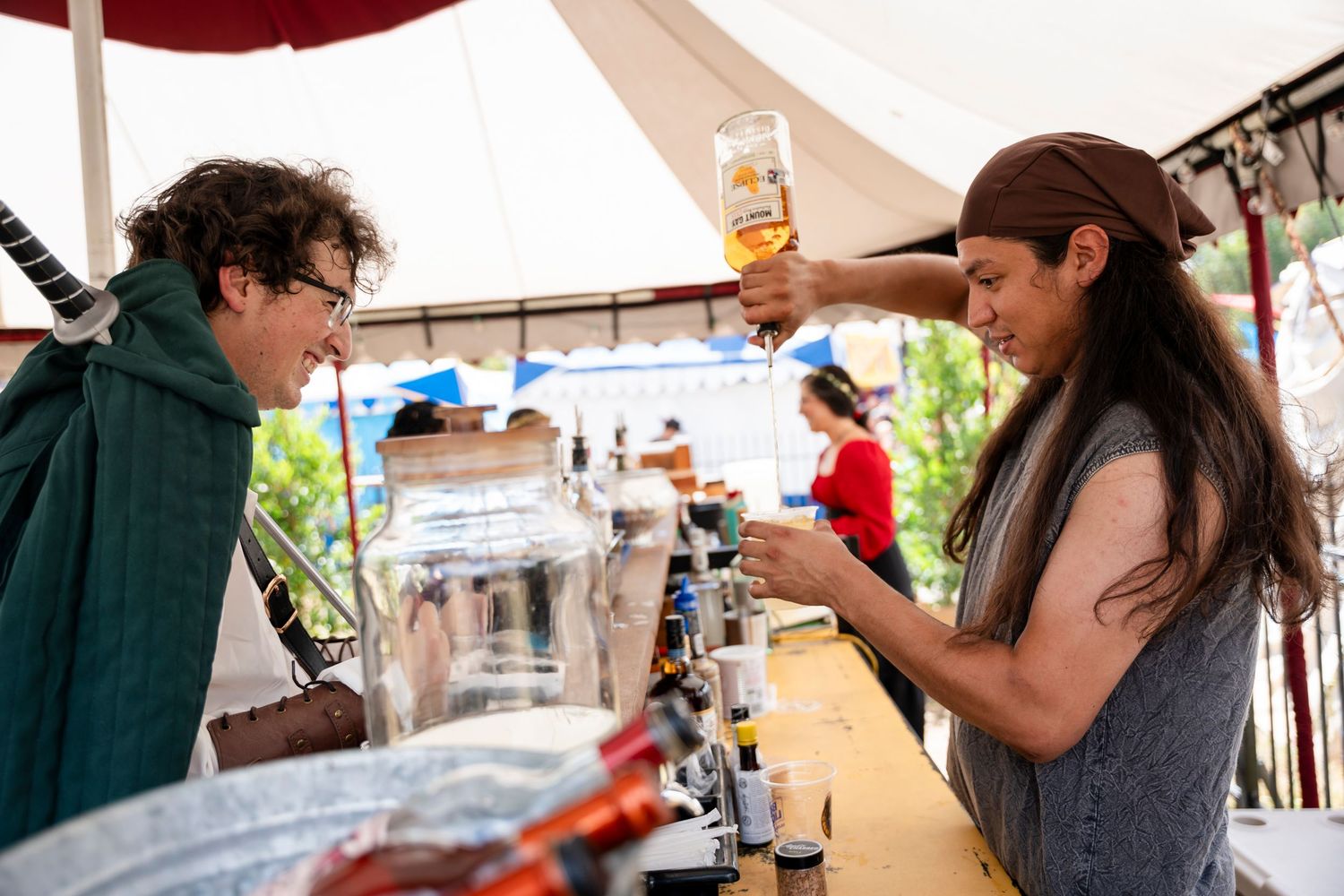 People enjoying drinks and socializing at an outdoor festival or market event under white tents.