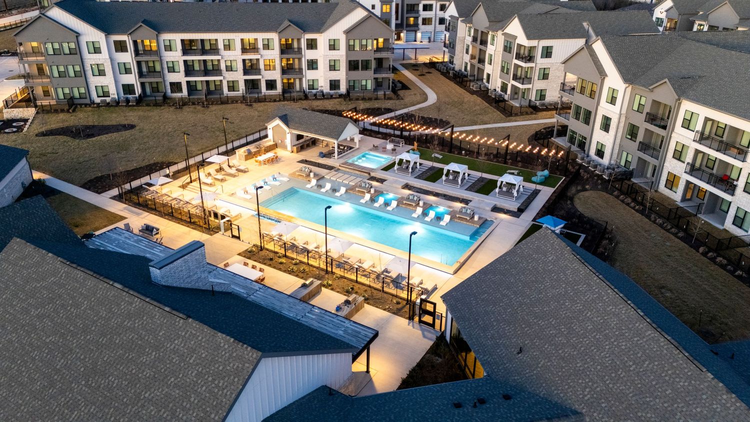 Aerial view of a modern apartment complex with illuminated swimming pool and deck area at dusk.