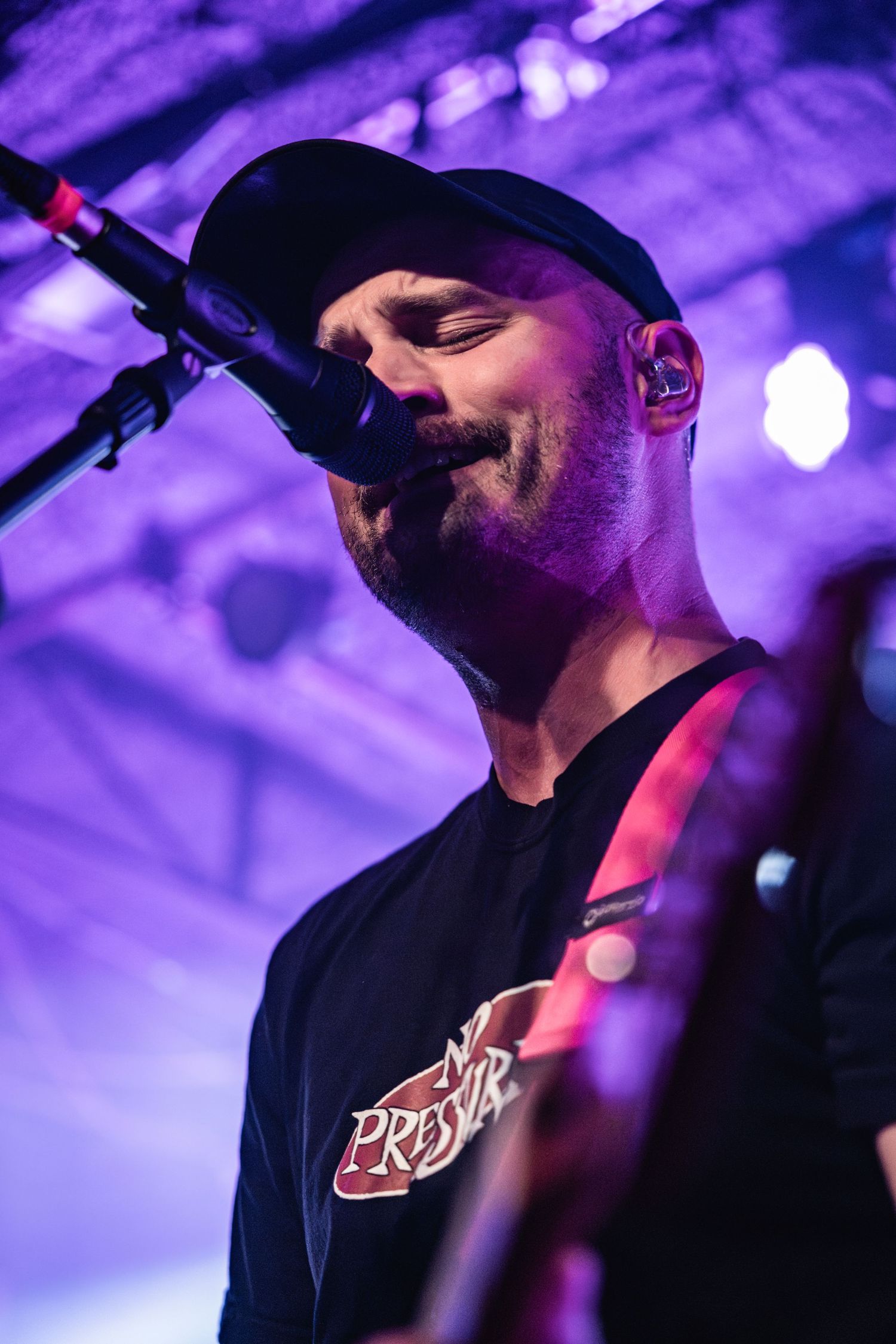Singer performing on stage in black baseball cap and t-shirt with purple stage lighting in background.