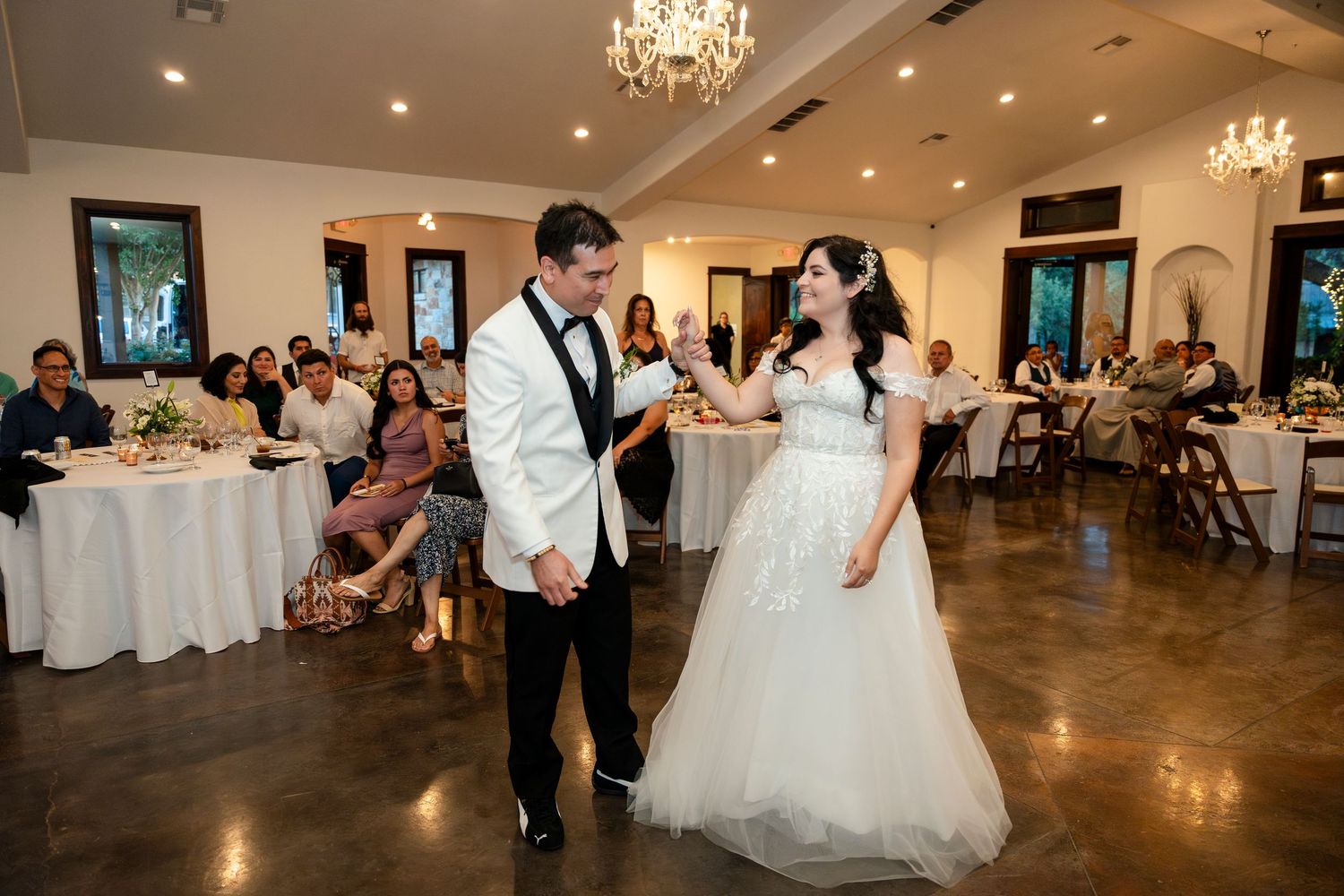 A bride and groom share their first dance together in an elegant ballroom with chandeliers and wedding guests watching.