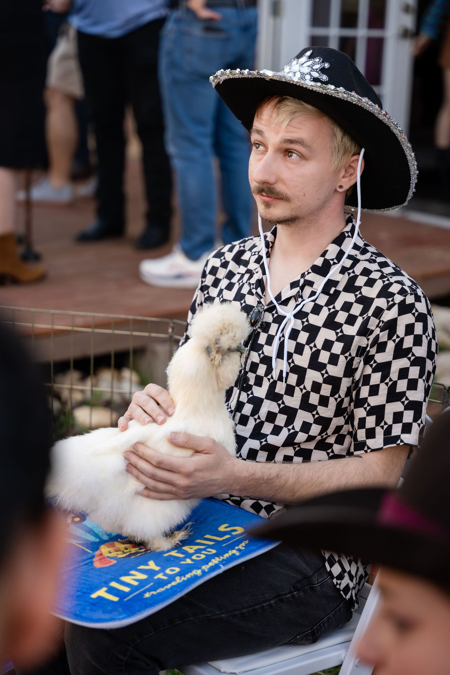 Person in black and white patterned shirt and decorated hat holding a small white pet animal with a blue sign nearby.