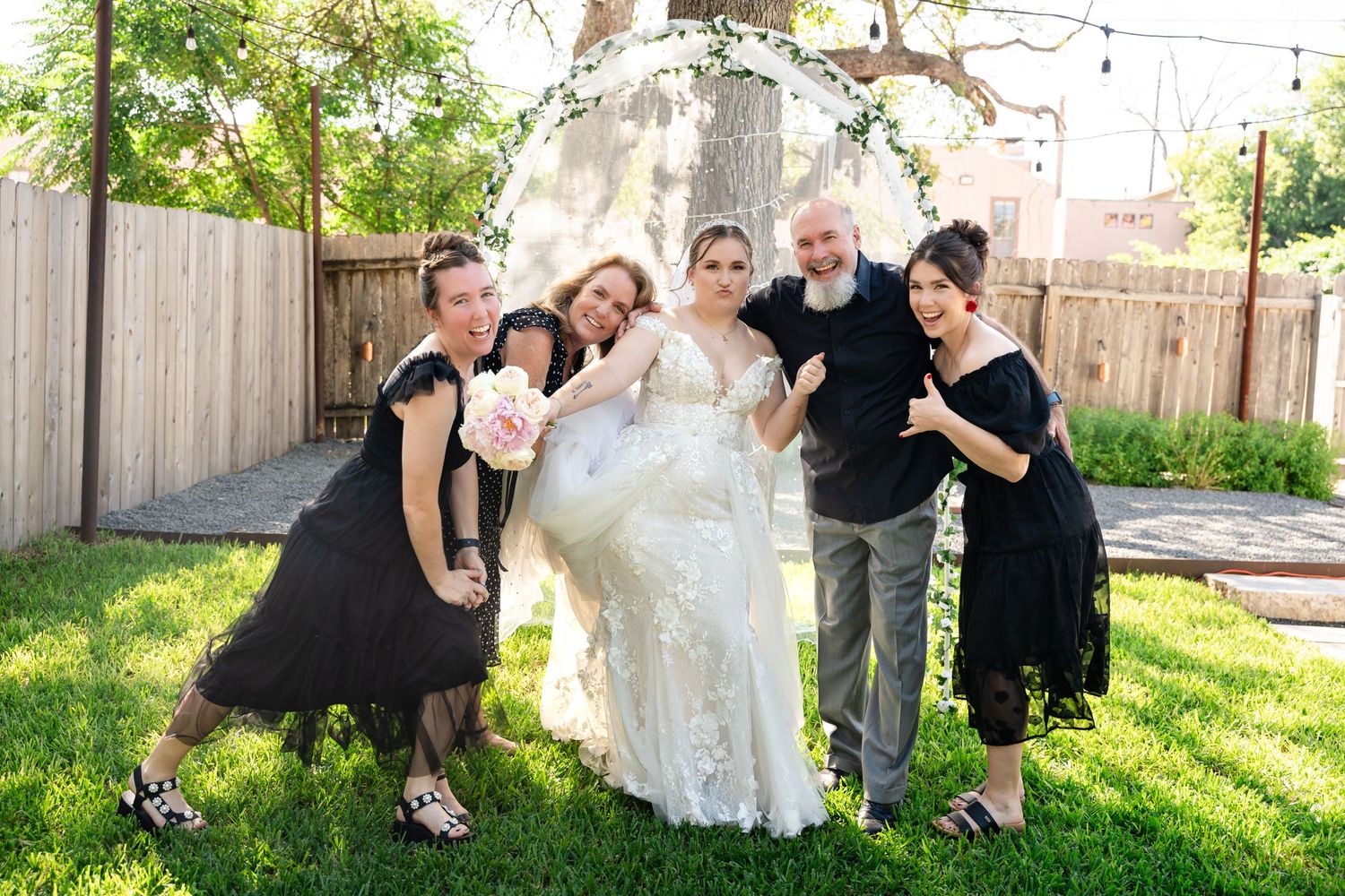 Wedding party poses playfully on grass with the bride in white gown surrounded by attendants in black.
