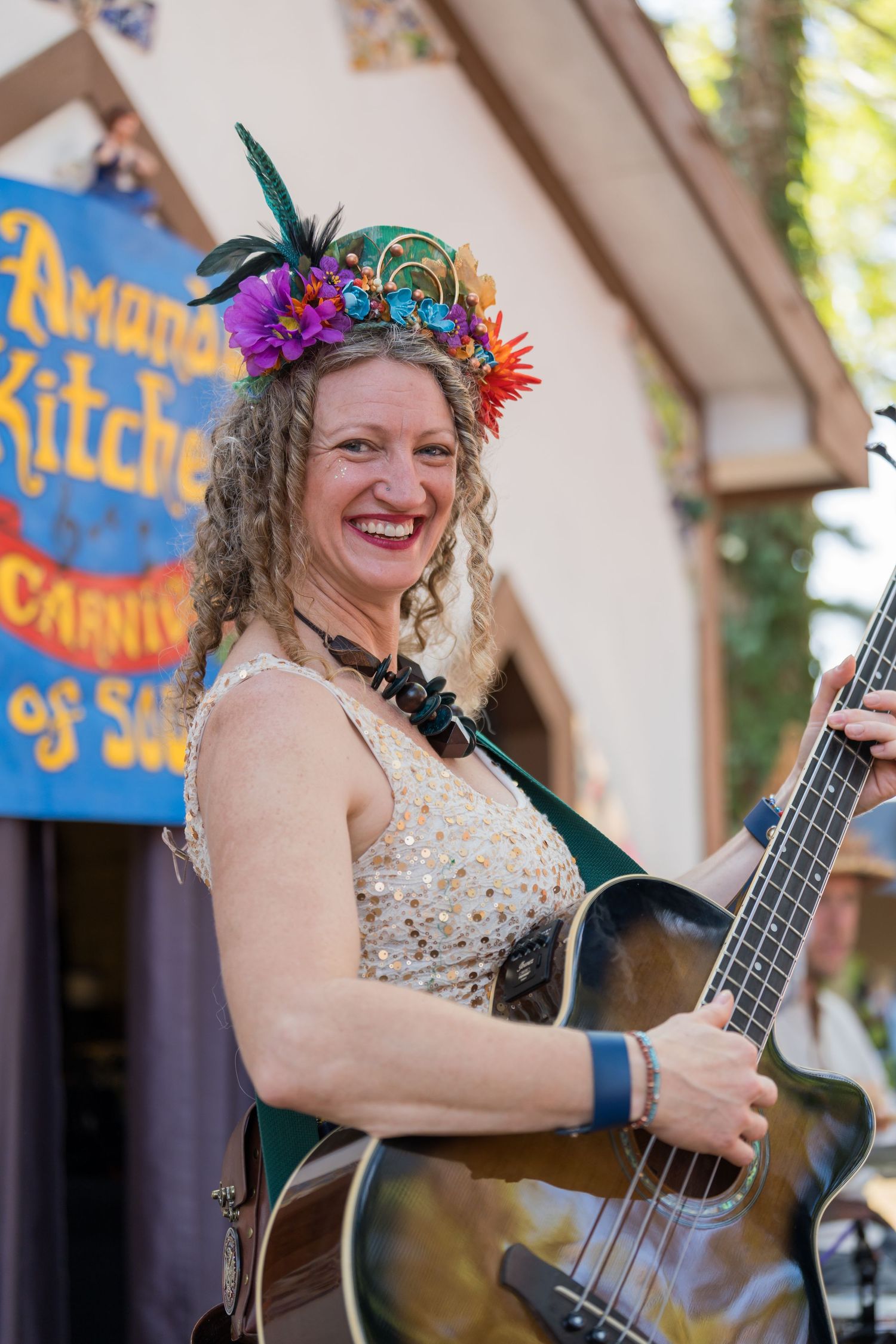 A performer wearing a colorful flower crown smiles while holding an acoustic guitar at a renaissance festival.