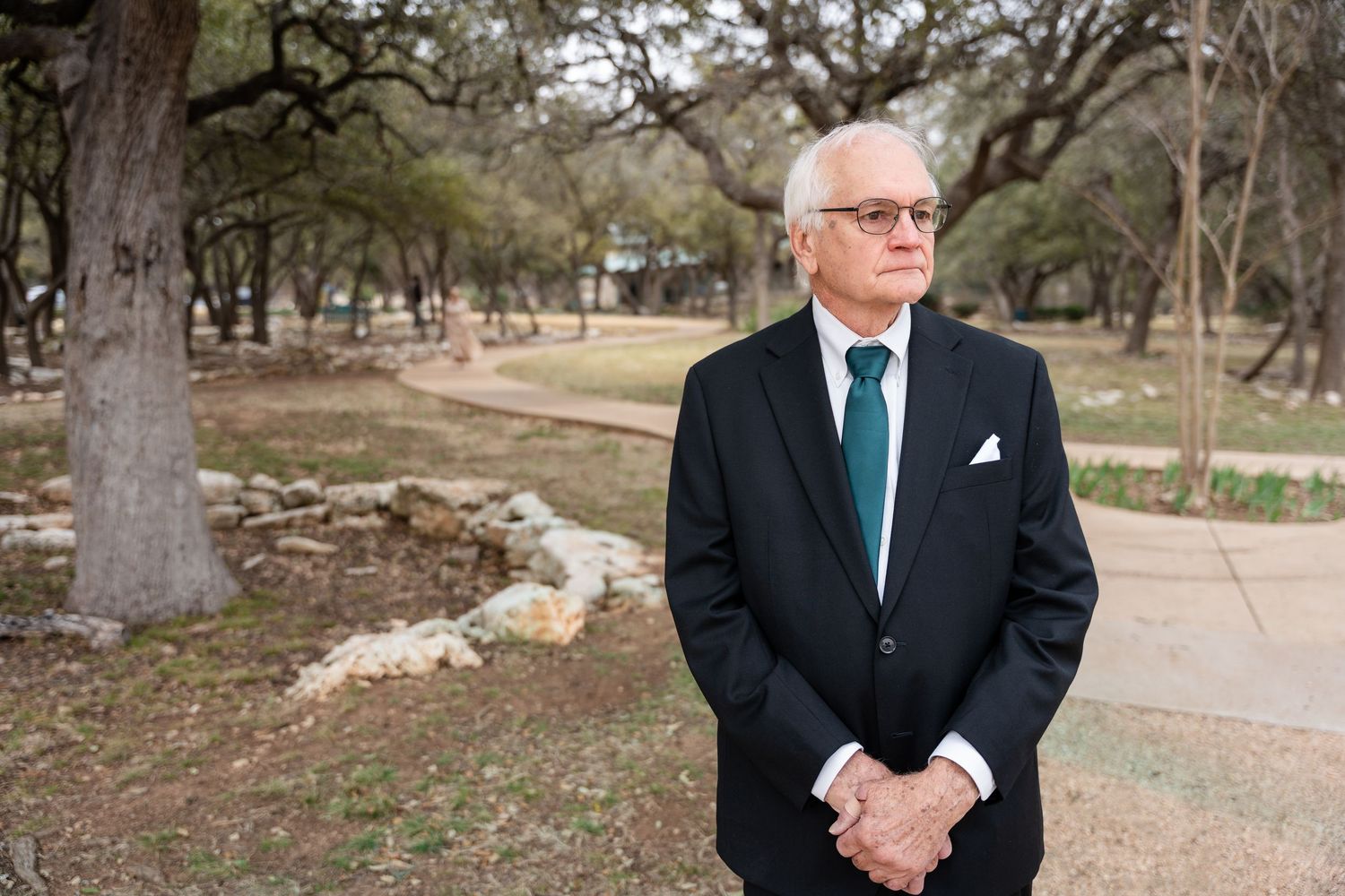 An elegantly dressed person in a dark suit with turquoise tie stands contemplatively in a natural outdoor park setting.