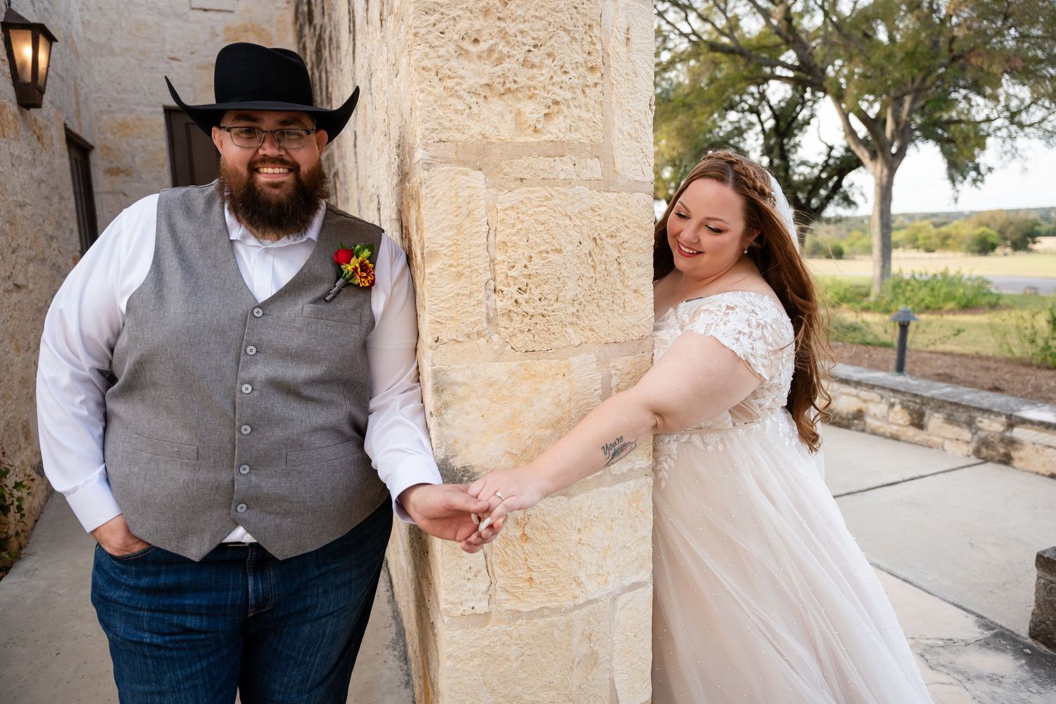 A couple in western-themed wedding attire pose playfully against a stone building exterior.