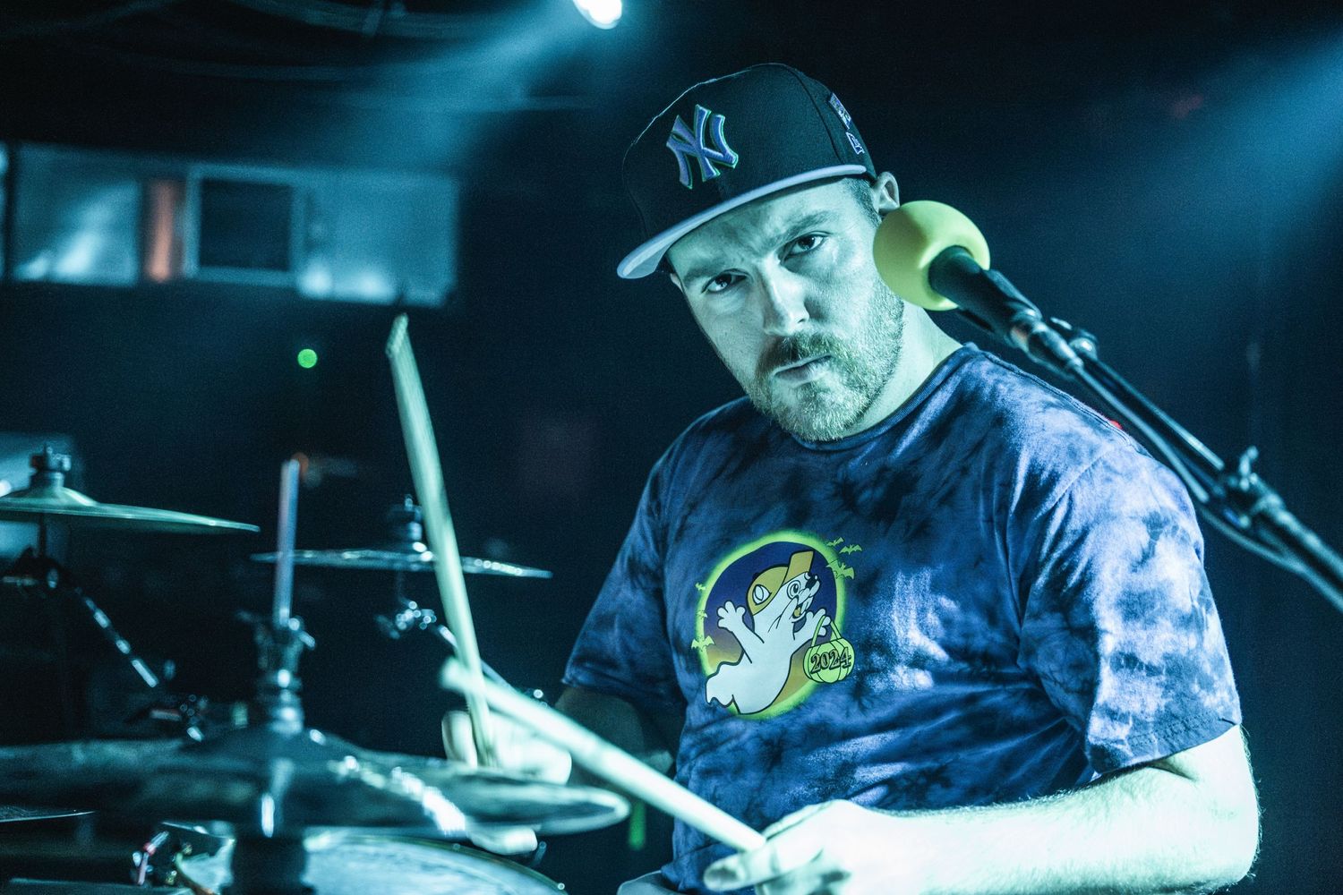 A drummer in a Yankees cap and tie-dye shirt performs under blue stage lighting.
