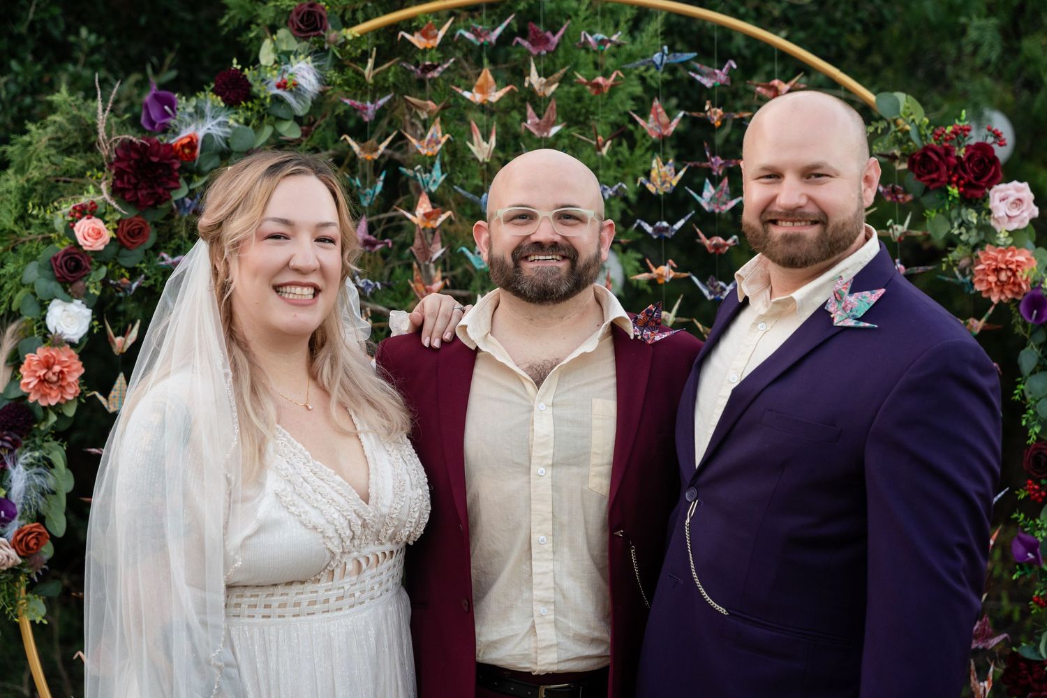 Wedding party poses in front of floral arch with origami crane backdrop during outdoor celebration.