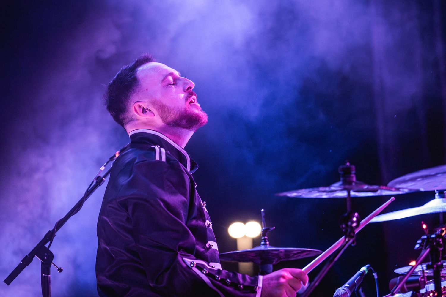Drummer performing on stage surrounded by dramatic blue and purple stage lighting effects.