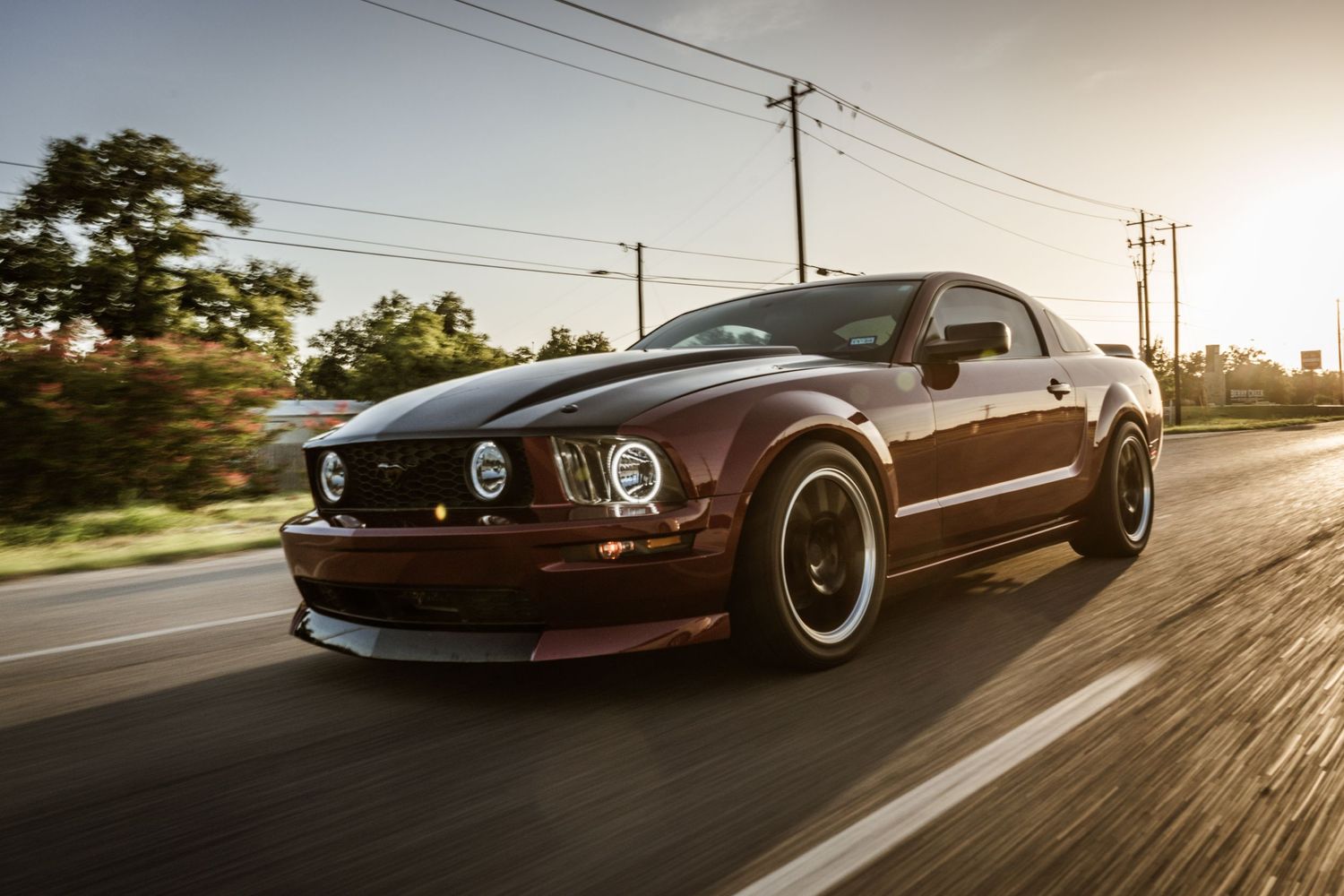 Burgundy Ford Mustang GT with black wheels speeding down a road during sunset.