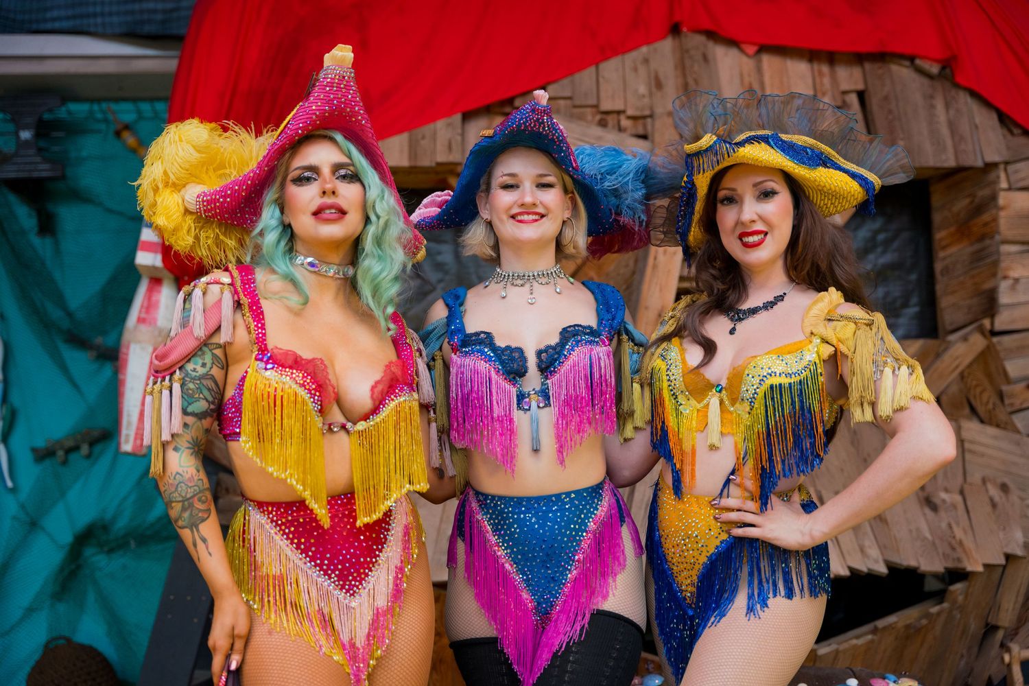 Three performers in colorful fringed burlesque costumes pose together against a red backdrop at a festival.