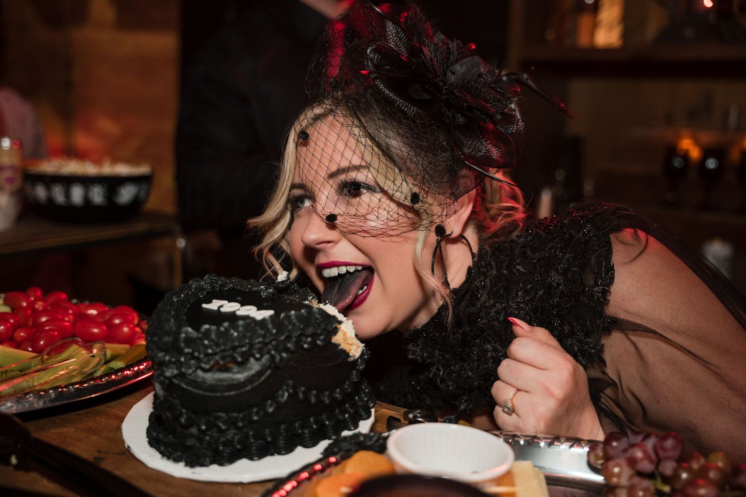 A person in vintage attire enthusiastically eating a black frosted cake at a party.