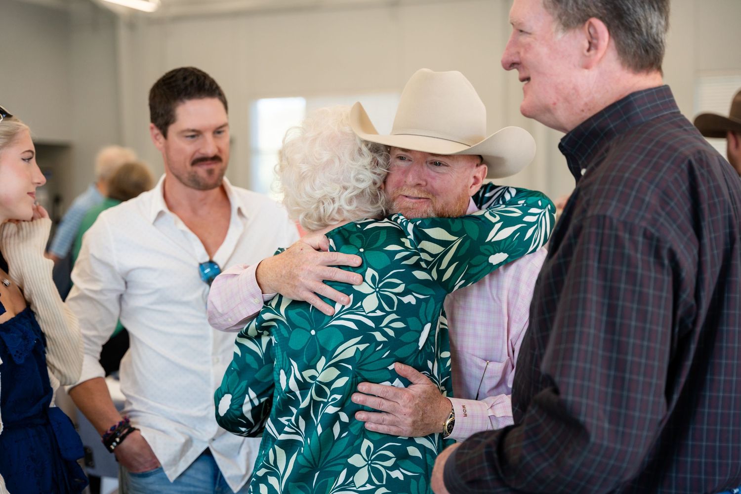 A group gathering with embraces, featuring someone in a white shirt and another in a green floral dress.