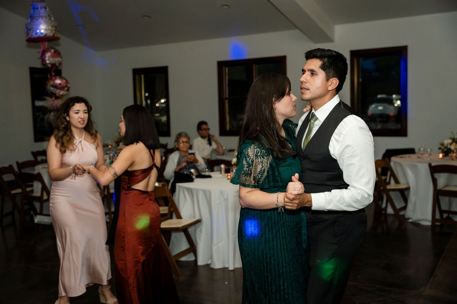 Couples dance at an evening reception with colorful blue lighting and round tables in the background.