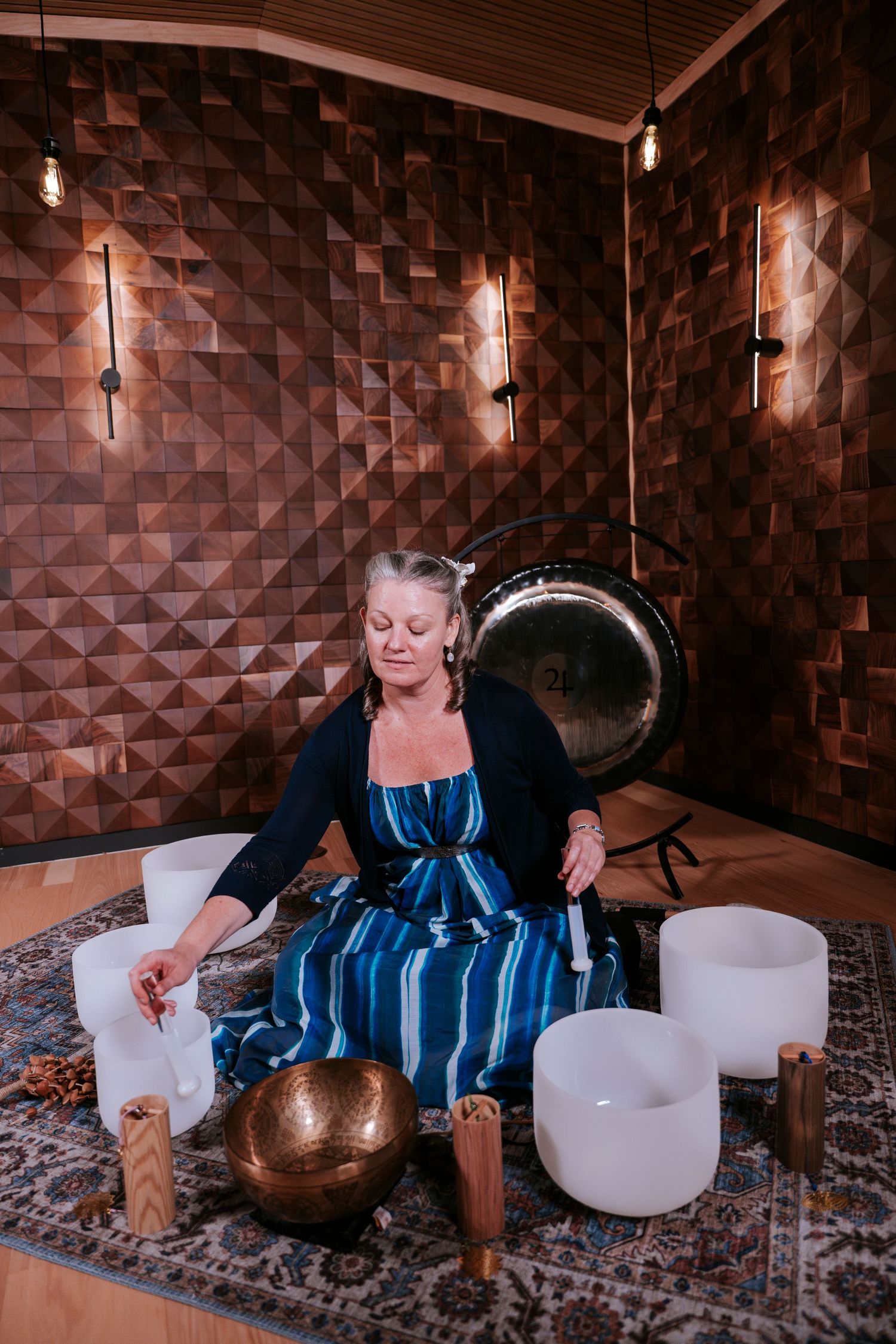 A person in blue dress sits on floor with crystal bowls, candles and meditation instruments in a wood-paneled room.