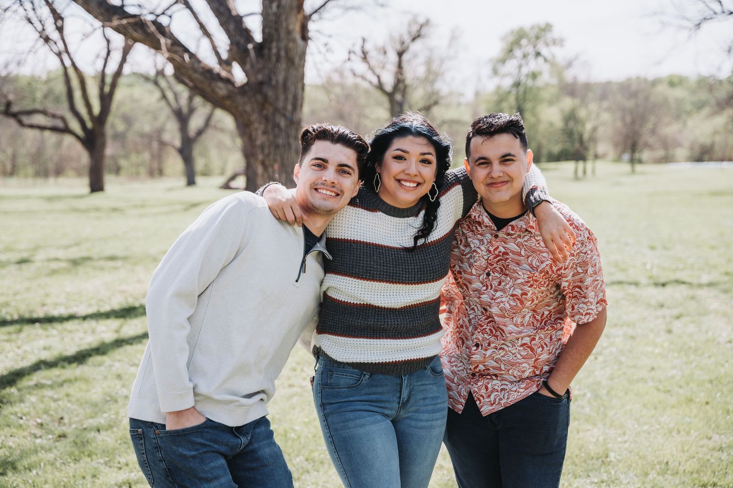 Three friends pose together outdoors on a sunny day with bare trees in background.