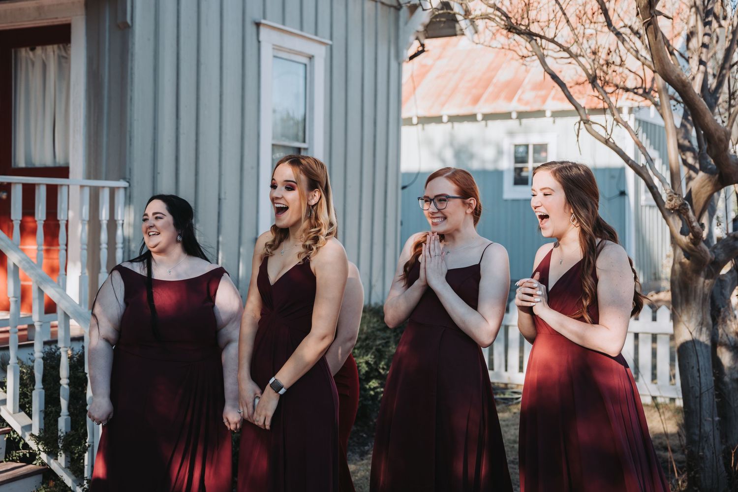 Four bridesmaids in burgundy dresses share a candid moment on a porch.