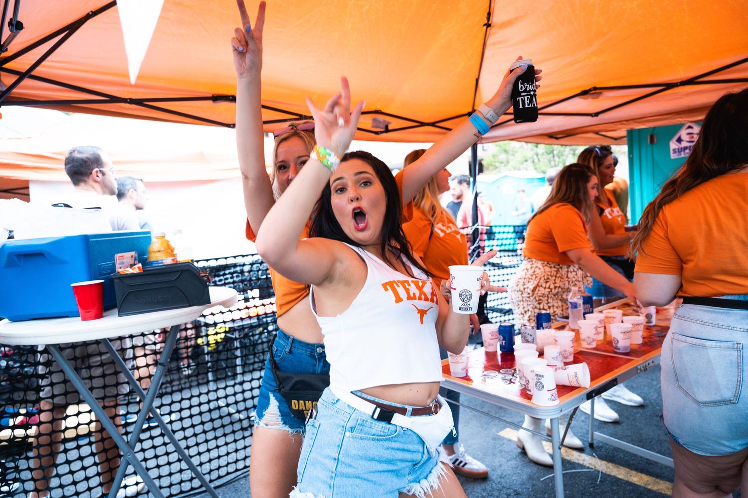 People celebrating enthusiastically under an orange tent at an outdoor party with drinks and snacks.