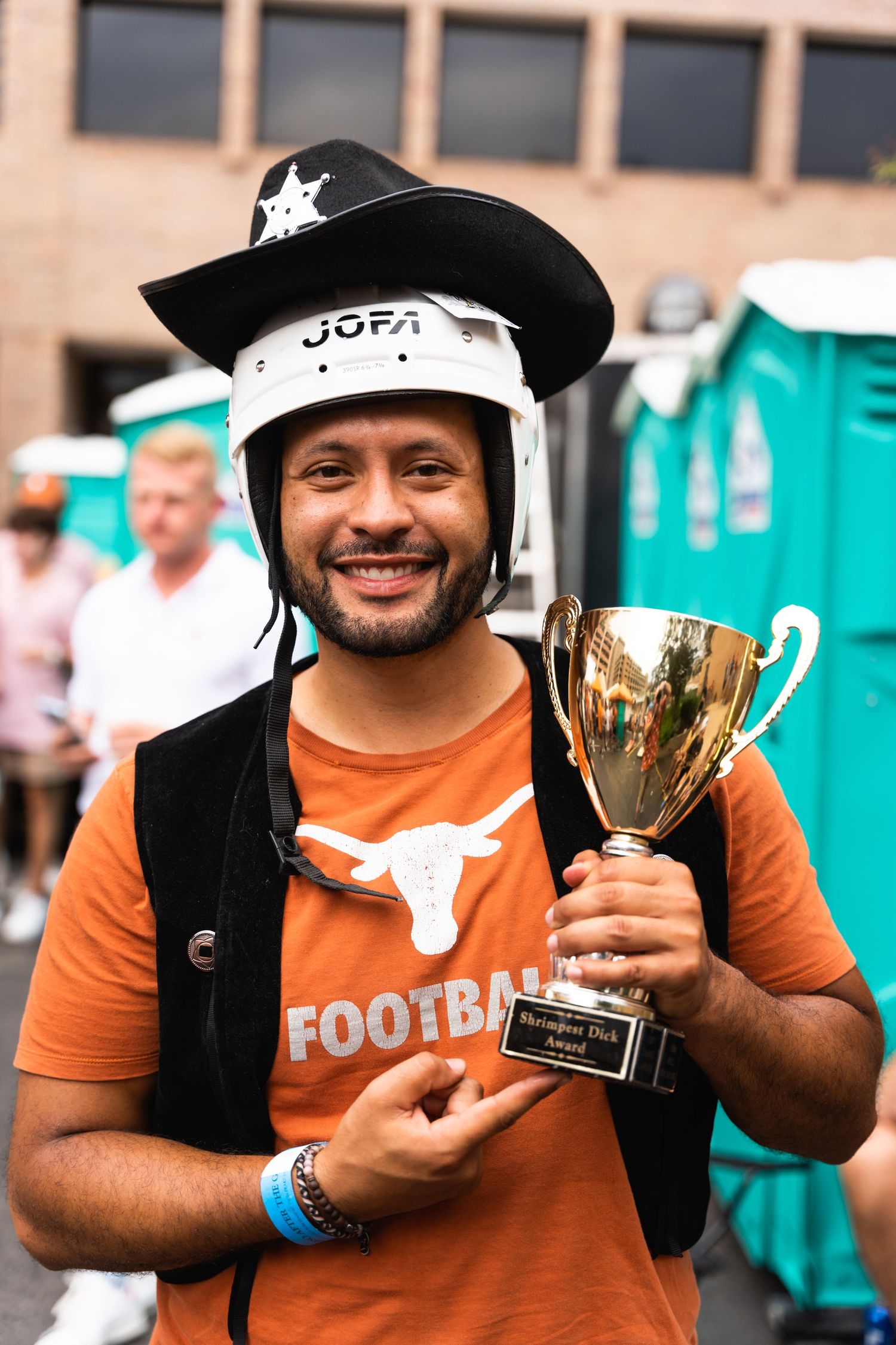 A sports fan wearing a Texas Football shirt and sheriff hat celebrates with a trophy at an outdoor event.