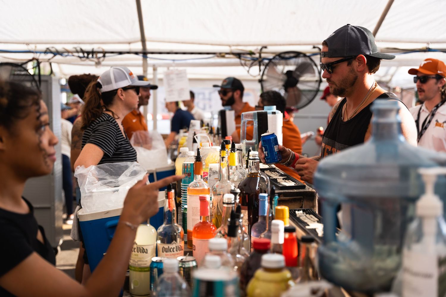 Busy outdoor bar setup with bartenders serving drinks under white tent.