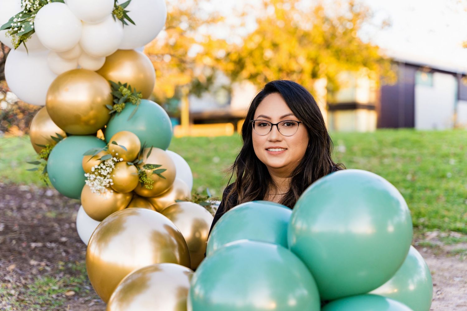 Person posing next to decorative balloon installation with gold and mint color scheme outdoors.