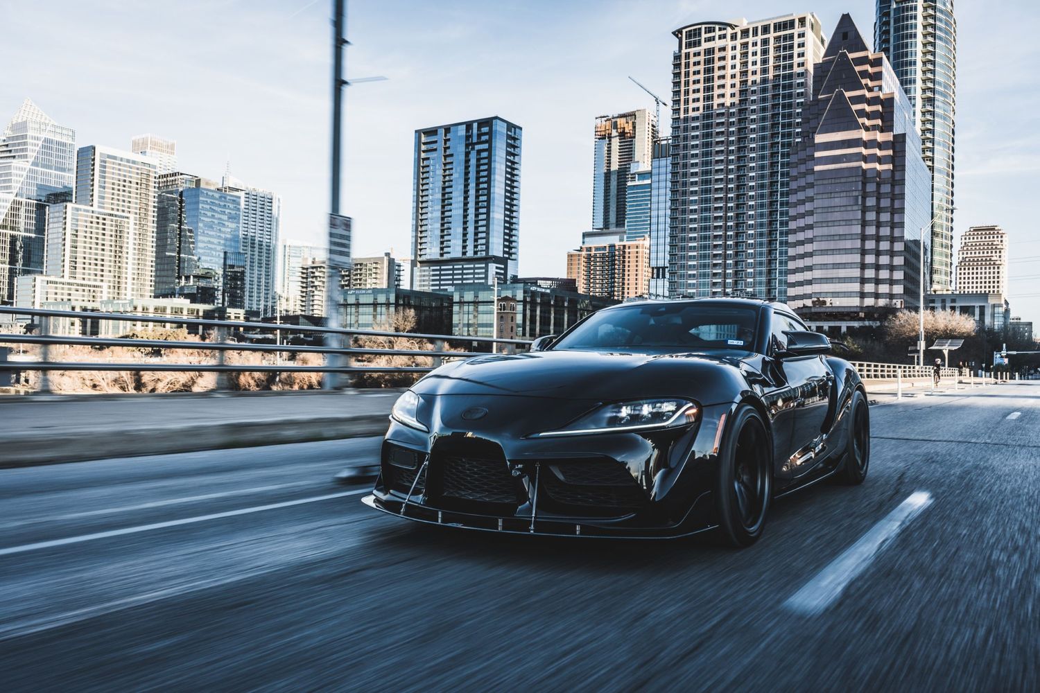 Modified black Toyota Supra driving on highway with modern Austin skyline in background.