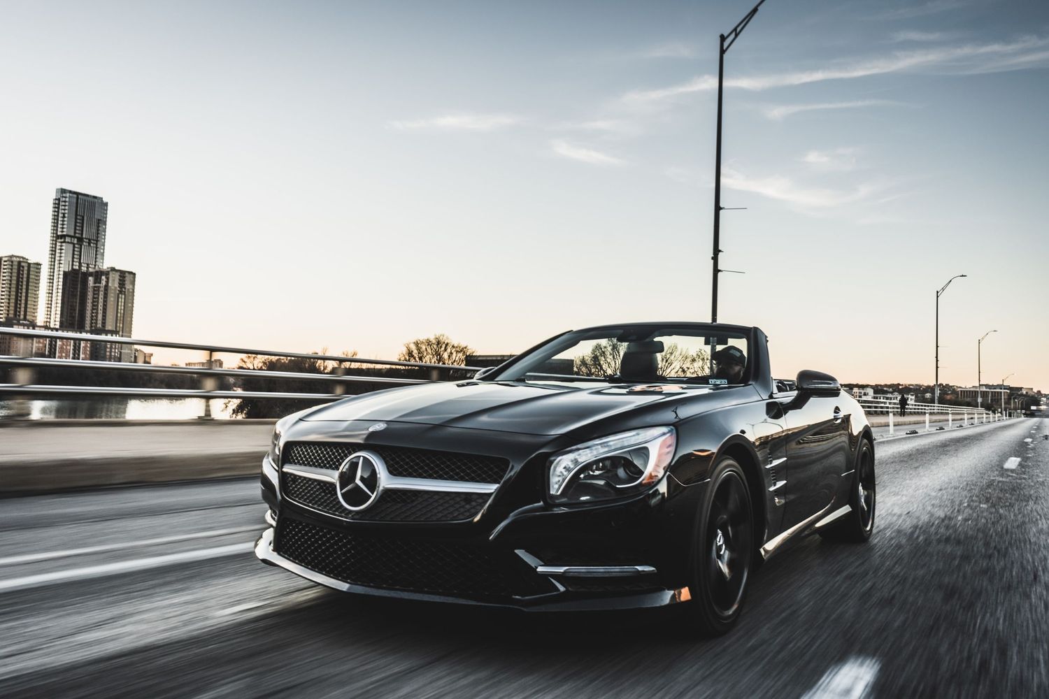 Black Mercedes-Benz convertible driving on urban bridge during sunset with city skyline in background.