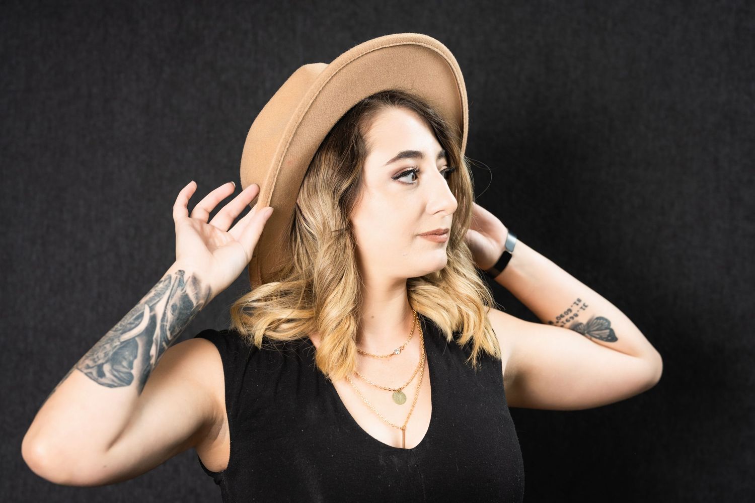 Artistic pose adjusting tan hat with visible tattoos and black sleeveless top.