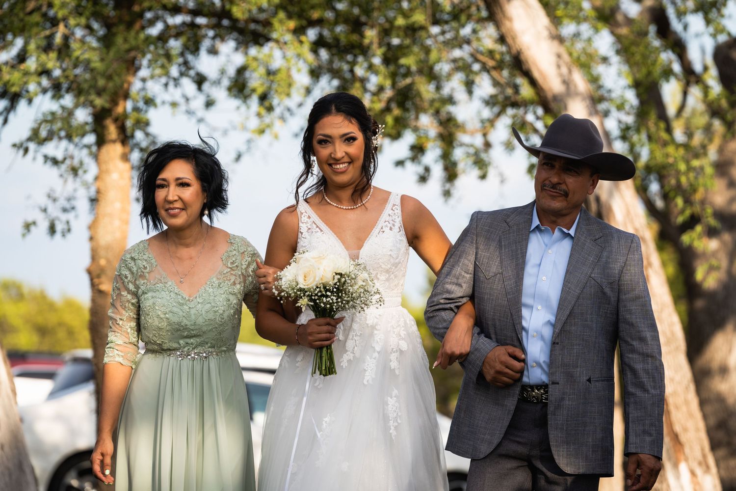 A bride in white walks between two wedding attendants at an outdoor ceremony.