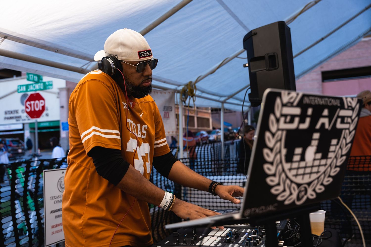 DJ performing at an outdoor event wearing an orange jersey behind professional equipment.