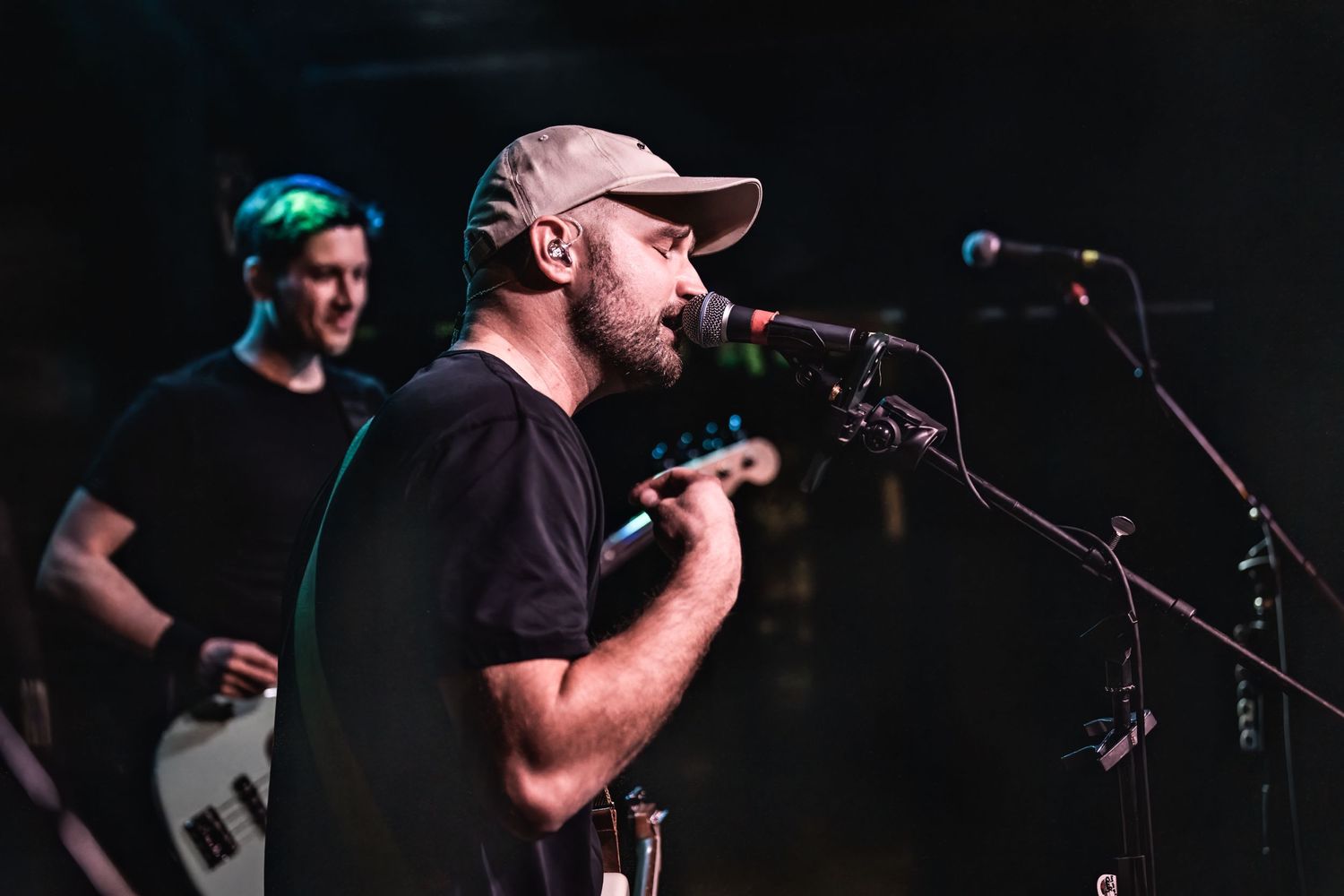 Vocalist in dark attire performs on stage with band member visible in background.